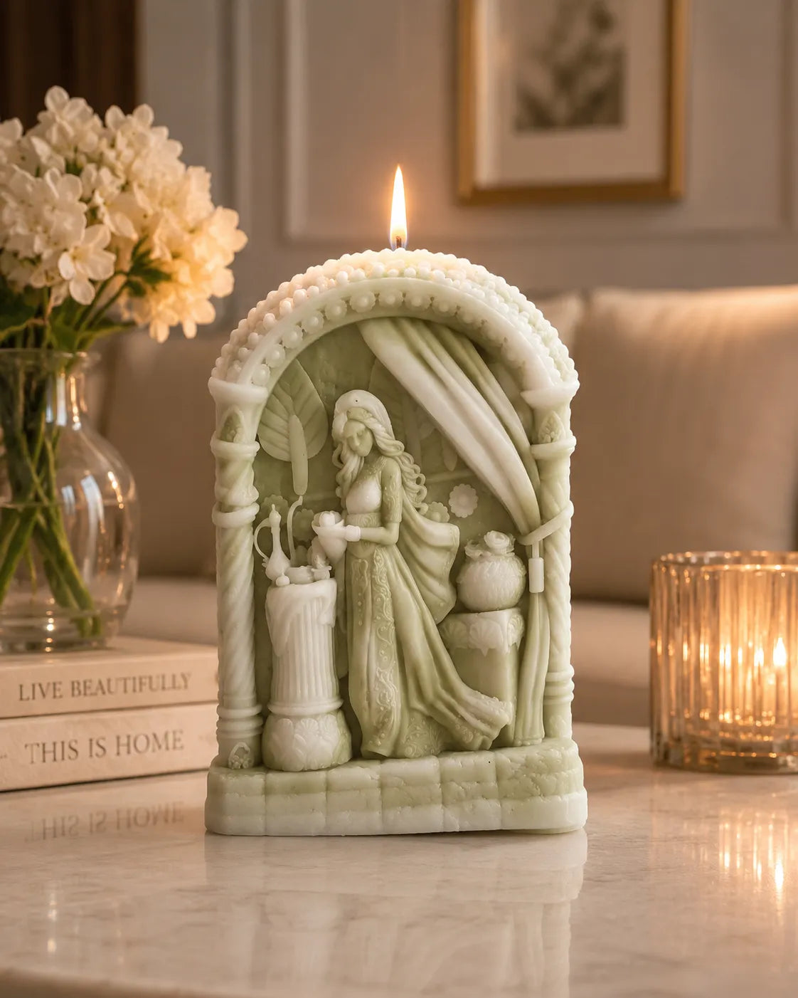 Decorative candle with a figurine of a woman in a dress, surrounded by flowers and books on a table.