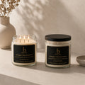 Two candles with black lids and labels on a neutral surface with a vase and plant in the background.