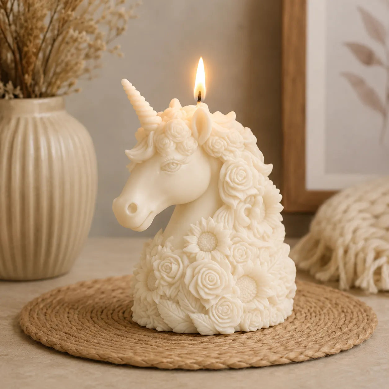 Unicorn-shaped candle with floral details on a woven mat, with a vase of dried flowers in the background.