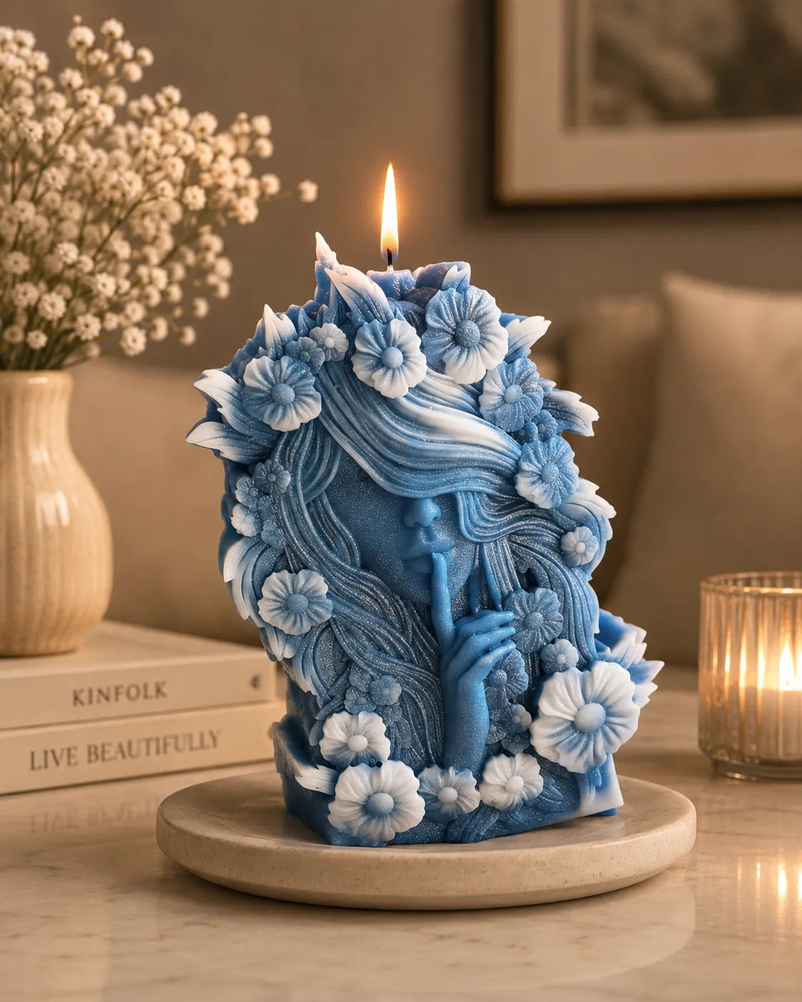 Decorative blue candle with floral patterns on a table with books and a vase in the background.